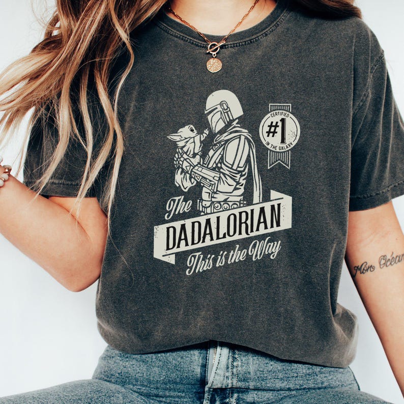 K&ouml;nnte beinhalten: Ein dunkelgraues T-Shirt mit einem Motiv eines Mandalorianer-Charakters, der ein Baby Yoda h&auml;lt. Der Text "The Dadalorian This is the Way" ist auf dem Shirt aufgedruckt. Das Motiv enth&auml;lt auch ein Abzeichen mit der Aufschrift "Certified #1 in the Galaxy".