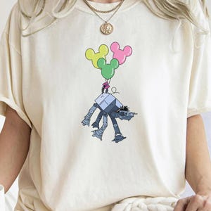 May include: A white t-shirt with a graphic of a robot holding onto three Mickey Mouse-shaped balloons. The balloons are pink, yellow, and green.