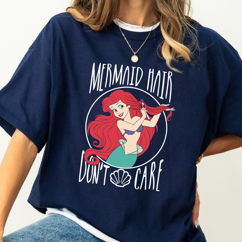 May include: A navy blue t-shirt with a graphic of Ariel from The Little Mermaid combing her hair. The text "Mermaid Hair Don't Care" is printed on the shirt.