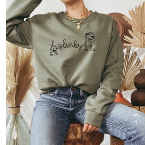 May include: A green sweatshirt with a black outline of Slinky Dog from the Toy Story franchise. The text "Slinky" is written in a cursive font above the dog.