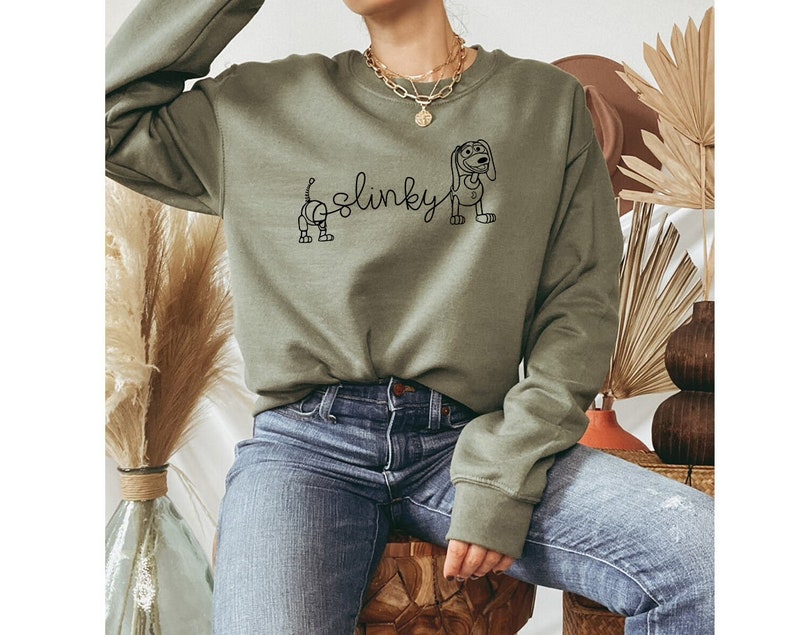 May include: A green sweatshirt with a black outline of Slinky Dog from the Toy Story franchise. The text "Slinky" is written in a cursive font above the dog.