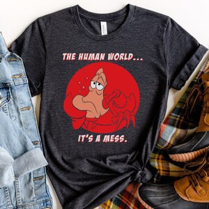 Op de afbeelding: Een donkergrijs T-shirt met een rode cirkel en een cartoonkrab met een droevige uitdrukking. De tekst "THE HUMAN WORLD... IT'S A MESS." is op het shirt gedrukt.
