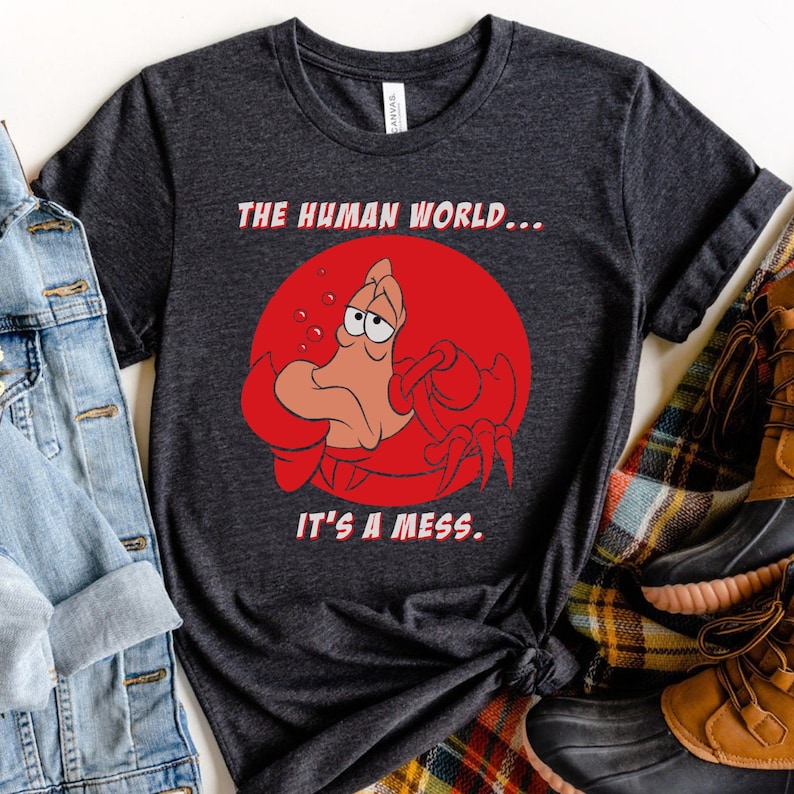 Op de afbeelding: Een donkergrijs T-shirt met een rode cirkel en een cartoonkrab met een droevige uitdrukking. De tekst "THE HUMAN WORLD... IT'S A MESS." is op het shirt gedrukt.