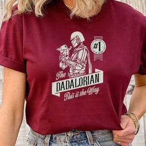 K&ouml;nnte beinhalten: Ein maroonfarbenes T-Shirt mit einer wei&szlig;en Grafik eines Mandalorianer-Charakters, der ein Baby Yoda h&auml;lt. Der Text auf dem Shirt lautet "The Dadalorian This is the Way" und "Certified #1 in the Galaxy".