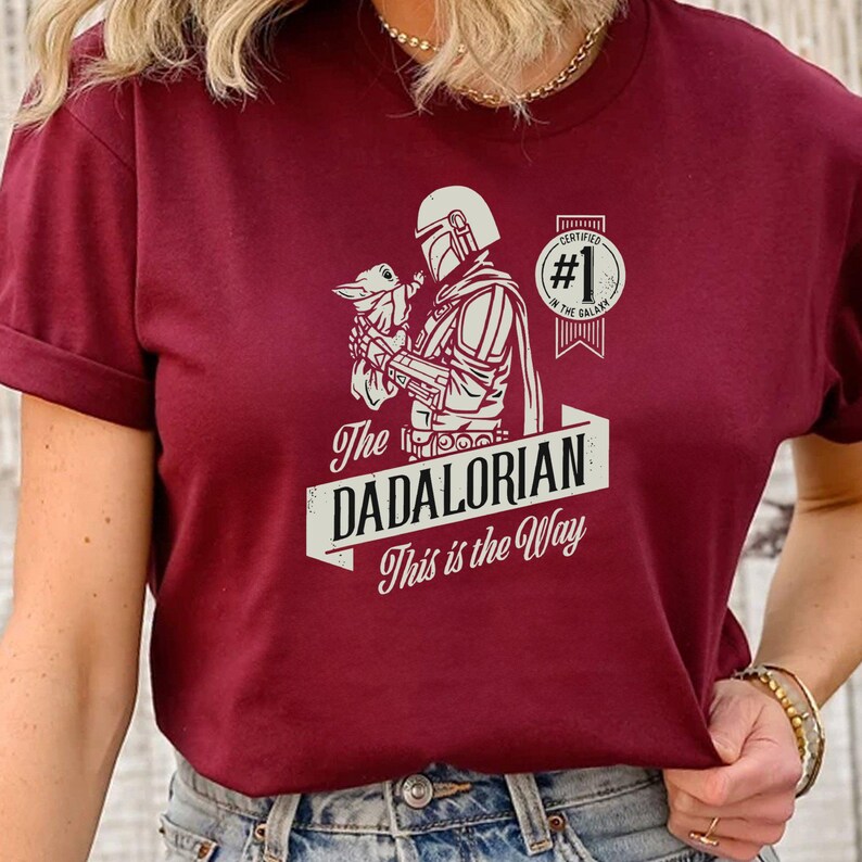 K&ouml;nnte beinhalten: Ein maroonfarbenes T-Shirt mit einer wei&szlig;en Grafik eines Mandalorianer-Charakters, der ein Baby Yoda h&auml;lt. Der Text auf dem Shirt lautet "The Dadalorian This is the Way" und "Certified #1 in the Galaxy".