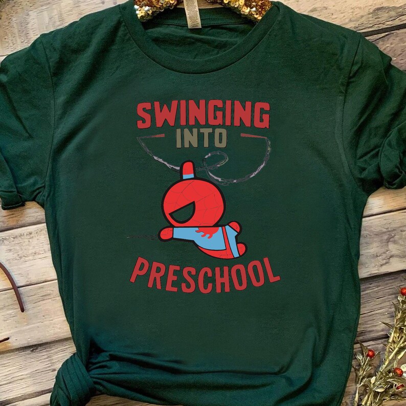 Puede incluir: Una camiseta verde oscuro con el texto "Swinging into Preschool" y una imagen de dibujos animados de un superh&eacute;roe balance&aacute;ndose en una cuerda.