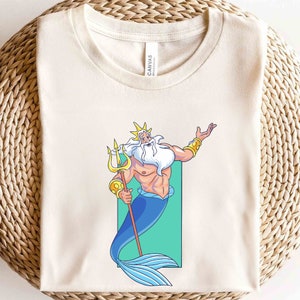 K&ouml;nnte beinhalten: Ein wei&szlig;es T-Shirt mit einem Cartoon-Bild von K&ouml;nig Triton aus Disneys Arielle, die Meerjungfrau. Er h&auml;lt seinen Dreizack und hat eine goldene Krone und einen Bart. Der Hintergrund ist hellgr&uuml;n.