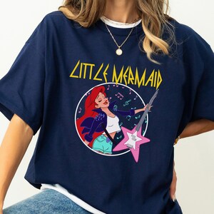 May include: A navy blue t-shirt with a graphic of Ariel from The Little Mermaid playing an electric guitar. The text "Little Mermaid" is printed in yellow above the graphic.