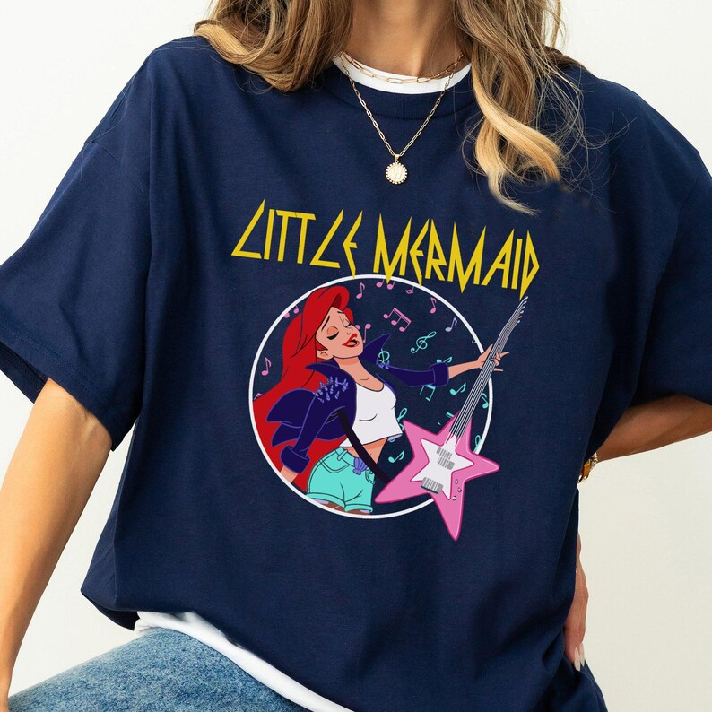 May include: A navy blue t-shirt with a graphic of Ariel from The Little Mermaid playing an electric guitar. The text "Little Mermaid" is printed in yellow above the graphic.