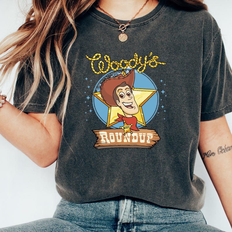 Peut inclure: Un t-shirt gris fonc&eacute; avec un graphisme vintage de Woody de Toy Story. Le graphisme repr&eacute;sente le visage de Woody, une &eacute;toile et le texte "Woody's Roundup".