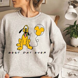 K&ouml;nnte beinhalten: Ein graues Sweatshirt mit einem Cartoon-Bild von Pluto, der einen gelben Ballon mit einem Mickey-Mouse-Ohren-Design h&auml;lt. Der Text "BEST DAY EVER" ist unter dem Bild aufgedruckt.