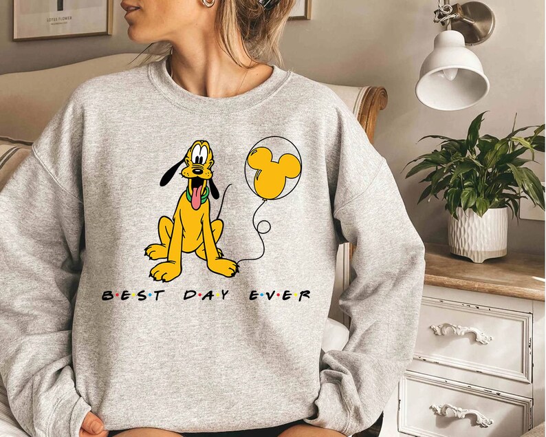K&ouml;nnte beinhalten: Ein graues Sweatshirt mit einem Cartoon-Bild von Pluto, der einen gelben Ballon mit einem Mickey-Mouse-Ohren-Design h&auml;lt. Der Text "BEST DAY EVER" ist unter dem Bild aufgedruckt.