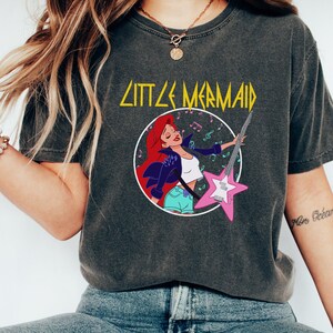 May include: A dark grey t-shirt with a graphic print of Ariel from The Little Mermaid playing an electric guitar. The text "Little Mermaid" is printed in yellow above the graphic.