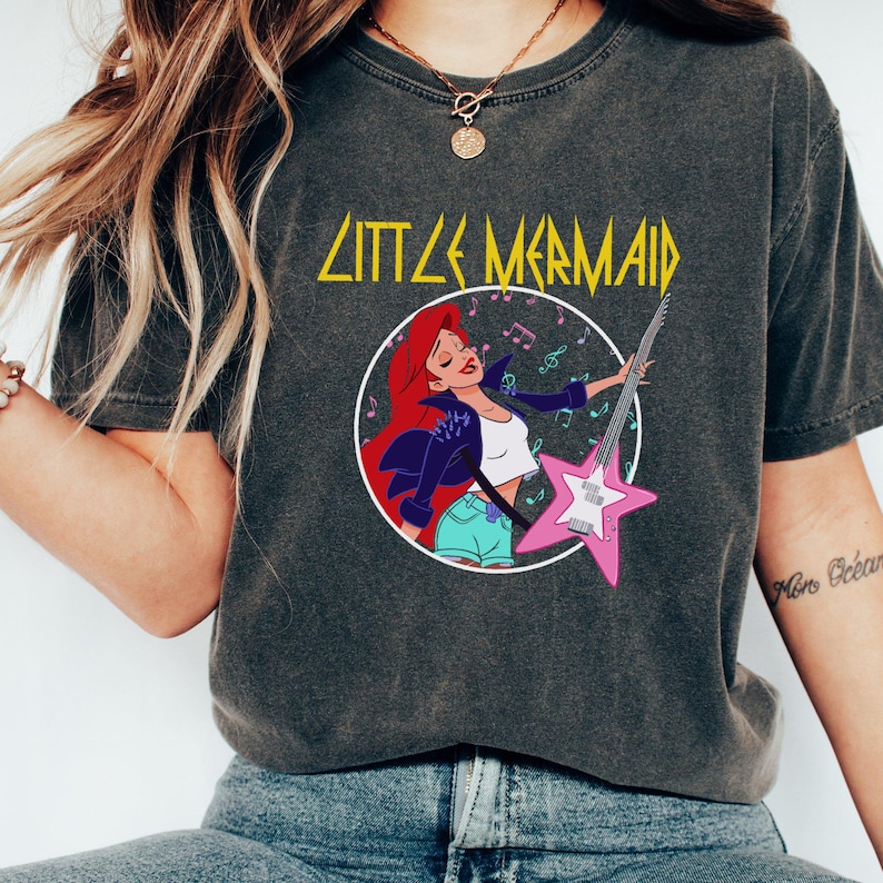 May include: A dark grey t-shirt with a graphic print of Ariel from The Little Mermaid playing an electric guitar. The text "Little Mermaid" is printed in yellow above the graphic.