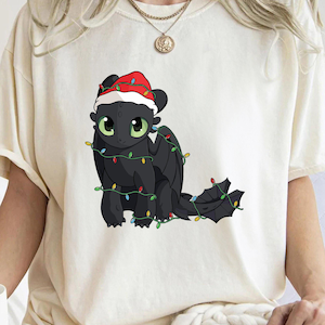 Peut inclure: Un t-shirt blanc avec un dragon de dessin animé noir portant un bonnet de Père Noël rouge et enveloppé de lumières de Noël colorées.