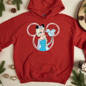 Puede incluir: Sudadera roja con capucha con un contorno blanco de las orejas de Mickey Mouse. Dentro de las orejas hay una imagen de dibujos animados de Elsa de Frozen sosteniendo un algod&oacute;n de az&uacute;car azul.