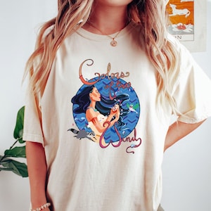 May include: A cream colored t-shirt with a graphic of Pocahontas from the Disney movie Pocahontas. The graphic features Pocahontas holding a seashell with the text "Colors of the Wind" in a stylized font.