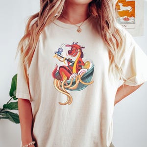 May include: A white t-shirt with a cartoon illustration of a red dragon eating noodles from a bowl. A small blue bug is perched on the rim of the bowl.