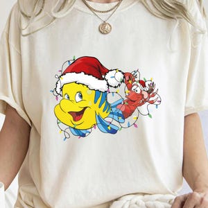Puede incluir: Una camiseta blanca con una imagen de dibujos animados de un pez amarillo con un gorro de Pap&aacute; Noel y una langosta roja sosteniendo luces de Navidad.