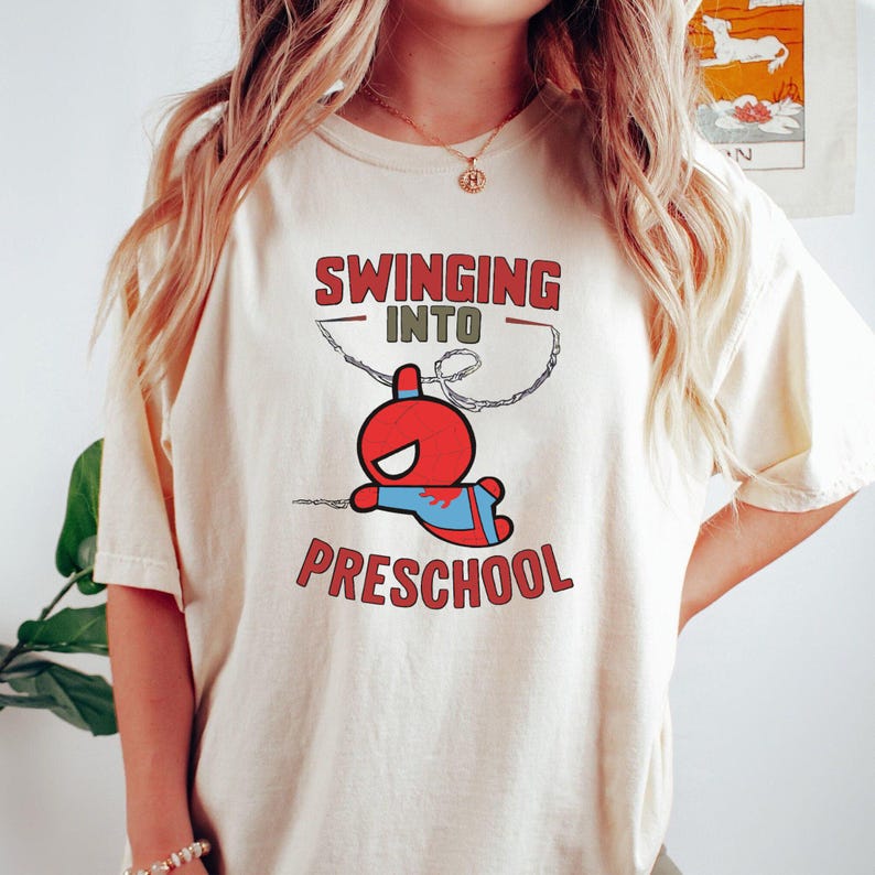 Puede incluir: Una camiseta blanca con el texto "Swinging into Preschool" y una imagen de dibujos animados de un superh&eacute;roe rojo y azul balance&aacute;ndose en una telara&ntilde;a.