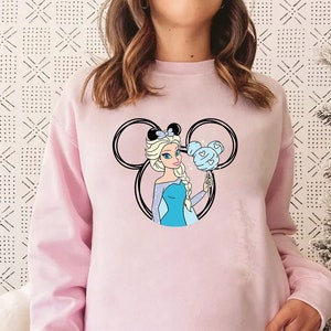 Puede incluir: Sudadera rosa con un gr&aacute;fico de una princesa Disney con cabello azul y un vestido azul que sostiene un algod&oacute;n de az&uacute;car azul en un palo. La princesa est&aacute; dentro de un contorno negro de las orejas de Mickey Mouse.