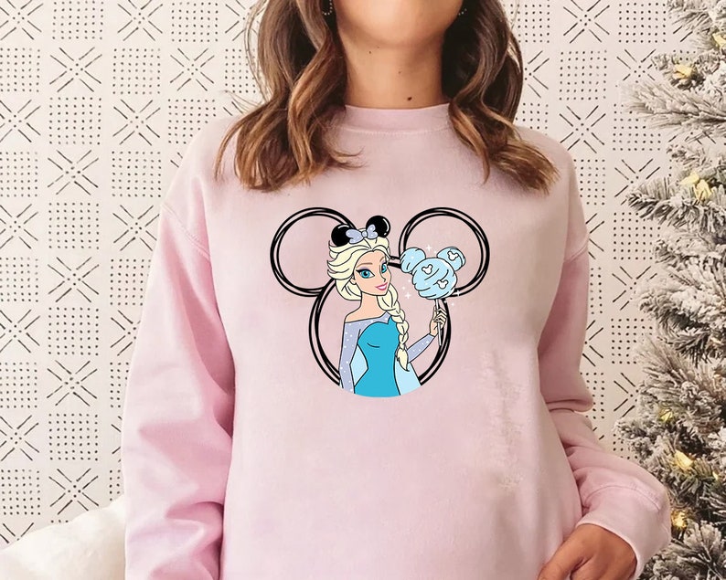 Puede incluir: Sudadera rosa con un gr&aacute;fico de una princesa Disney con cabello azul y un vestido azul que sostiene un algod&oacute;n de az&uacute;car azul en un palo. La princesa est&aacute; dentro de un contorno negro de las orejas de Mickey Mouse.