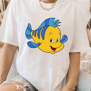 May include: White t-shirt with a cartoon yellow and blue fish with a smiling face.