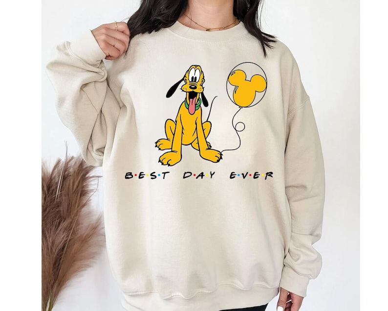 K&ouml;nnte beinhalten: Ein beiger Pullover mit einem Cartoon-Bild von Pluto, der einen roten Ballon mit Mickey-Maus-Ohren h&auml;lt. Der Text "BEST DAY EVER" ist unter dem Bild aufgedruckt.