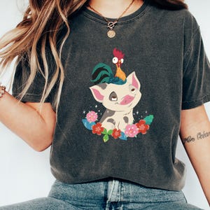 K&ouml;nnte beinhalten: Ein dunkelgraues T-Shirt mit einem Cartoon-Bild eines Schweins mit einem Hahn auf dem Kopf. Das Schwein ist wei&szlig; mit schwarzen Flecken und von rosa und gr&uuml;nen Blumen umgeben.