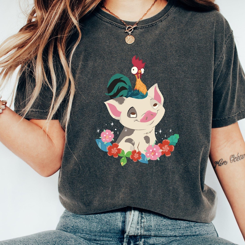 K&ouml;nnte beinhalten: Ein dunkelgraues T-Shirt mit einem Cartoon-Bild eines Schweins mit einem Hahn auf dem Kopf. Das Schwein ist wei&szlig; mit schwarzen Flecken und von rosa und gr&uuml;nen Blumen umgeben.