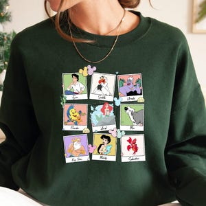 May include: Dark green sweatshirt with a grid of nine character portraits from The Little Mermaid. Characters include Eric, Scuttle, Ursula, Flounder, Ariel, Max, King Triton, Melody, and Sebastian. Each portrait is in a polaroid-style frame.