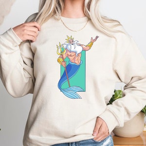 K&ouml;nnte beinhalten: Ein wei&szlig;er Pullover mit einem Cartoon-Bild von K&ouml;nig Triton aus Disneys Arielle, die Meerjungfrau. Er h&auml;lt einen Dreizack und hat einen blauen Schwanz.