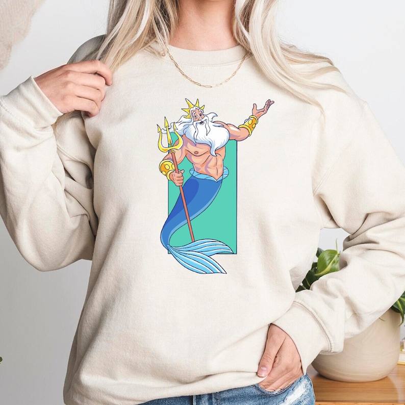 K&ouml;nnte beinhalten: Ein wei&szlig;er Pullover mit einem Cartoon-Bild von K&ouml;nig Triton aus Disneys Arielle, die Meerjungfrau. Er h&auml;lt einen Dreizack und hat einen blauen Schwanz.