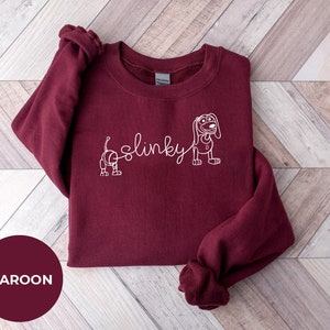 May include: Maroon sweatshirt with a white outline of Slinky Dog from Toy Story. The text "Slinky" is written in cursive.