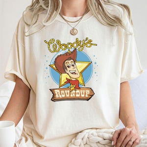 Peut inclure: Un t-shirt blanc avec une image de dessin anim&eacute; de Woody de Toy Story. L'image repr&eacute;sente Woody portant son chapeau de cowboy et son gilet embl&eacute;matiques, tenant un badge de sh&eacute;rif. Le texte "Woody's Roundup" est imprim&eacute; sous l'image.