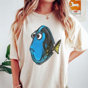 May include: A cream-colored t-shirt with a cartoon image of Dory, the blue tang fish from the movie Finding Nemo. Dory is looking to the left with her mouth open and her fin is extended.