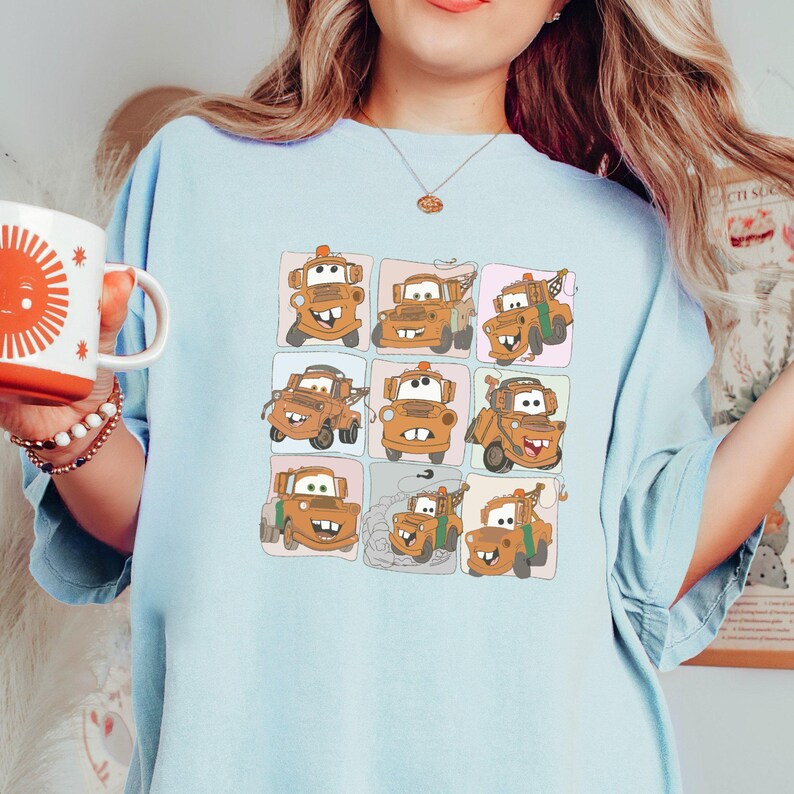 Puede incluir: Una camiseta azul claro con una imagen de dibujos animados de Mater de la pel&iacute;cula Cars de Disney Pixar. La imagen muestra a Mater en nueve poses diferentes, cada una en un cuadrado separado. Los cuadrados est&aacute;n dispuestos en una cuadr&iacute;cula de 3x3.