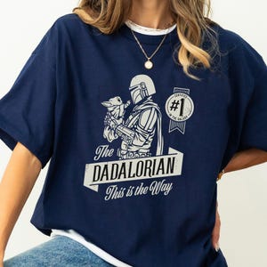 K&ouml;nnte beinhalten: Ein marineblaues T-Shirt mit einer wei&szlig;en Grafik eines Mandalorianer-Charakters, der ein Baby Yoda h&auml;lt. Die Grafik enth&auml;lt den Text "The Dadalorian This is the Way" und ein Abzeichen mit der Aufschrift "Certified #1 in the Galaxy".