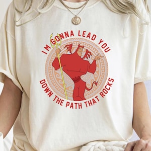 May include: A white t-shirt with a red cartoon devil character holding a golden trident. The text on the shirt reads "I'm gonna lead you down the path that rocks".