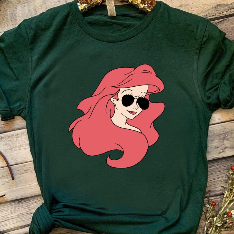 May include: A dark green t-shirt with a cartoon image of Ariel from The Little Mermaid wearing sunglasses. The image is in pink and black.