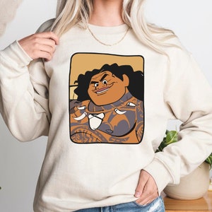 May include: A cream coloured sweatshirt featuring a cartoon image of a Polynesian man with tattoos and a mischievous grin. The image is framed in a brown rectangle.