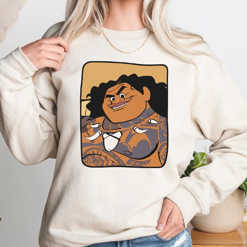 May include: A cream coloured sweatshirt featuring a cartoon image of a Polynesian man with tattoos and a mischievous grin. The image is framed in a brown rectangle.