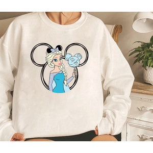 Puede incluir: Sudadera blanca con una ilustraci&oacute;n en blanco y negro de un personaje de dibujos animados con cabello azul y un vestido azul que sostiene un globo azul y blanco con forma de Mickey Mouse. El personaje lleva una corona.