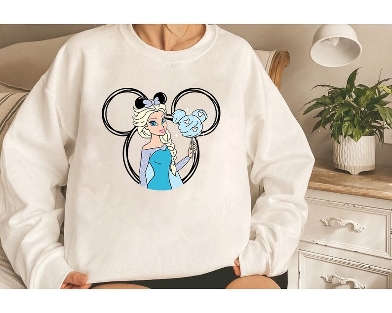 Puede incluir: Sudadera blanca con una ilustraci&oacute;n en blanco y negro de un personaje de dibujos animados con cabello azul y un vestido azul que sostiene un globo azul y blanco con forma de Mickey Mouse. El personaje lleva una corona.