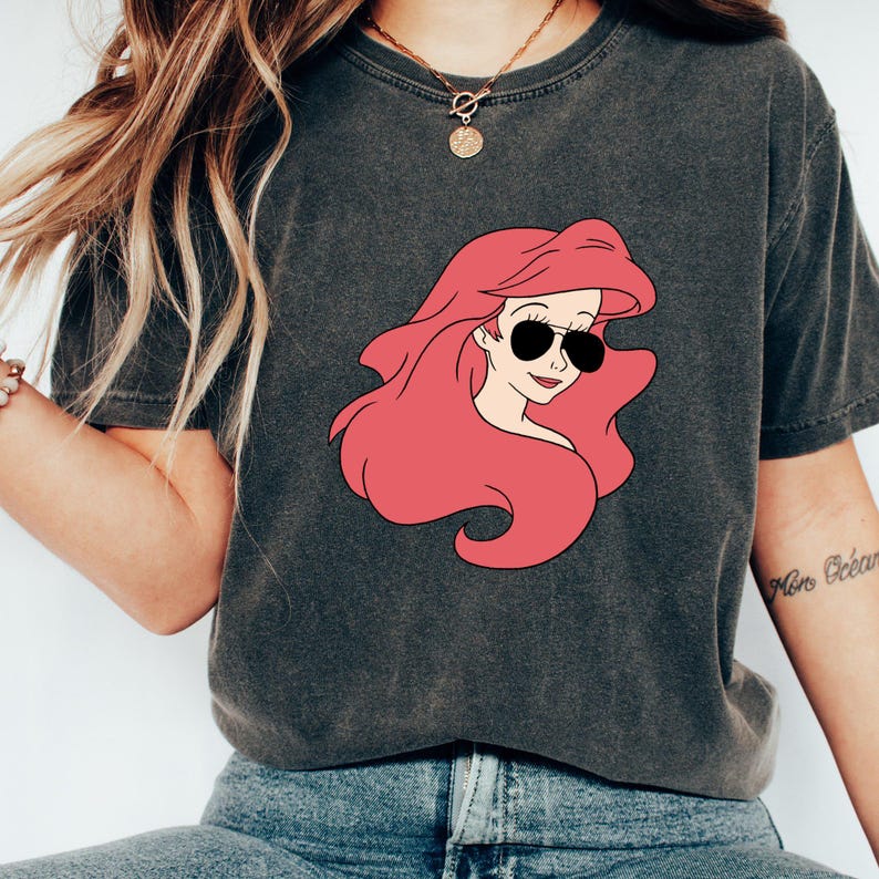 May include: A dark grey t-shirt with a cartoon image of Ariel from The Little Mermaid wearing sunglasses. The image is in pink and black.