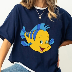 May include: A navy blue t-shirt with a cartoon image of Flounder, a yellow fish with blue fins, from the Disney movie The Little Mermaid.