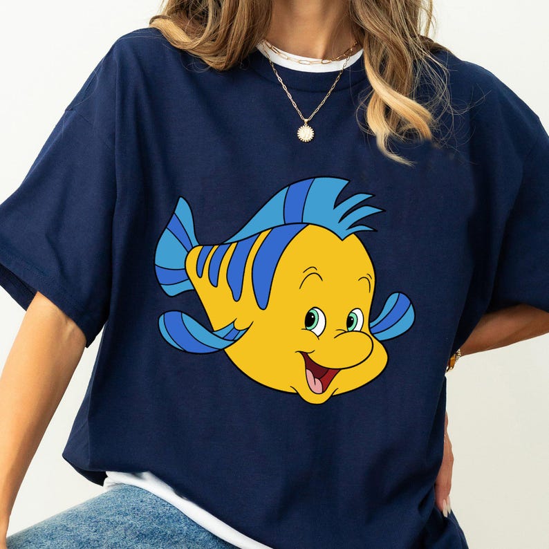 May include: A navy blue t-shirt with a cartoon image of Flounder, a yellow fish with blue fins, from the Disney movie The Little Mermaid.