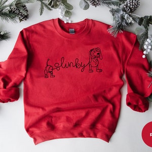 May include: Red sweatshirt with a black outline of Slinky Dog from Toy Story. The text "Slinky" is written in a cursive font.
