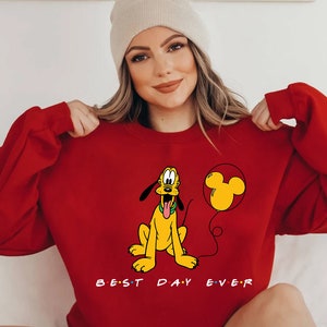 K&ouml;nnte beinhalten: Roter Pullover mit einem Cartoon-Bild von Pluto, der einen Mickey-Maus-Ballon h&auml;lt. Der Text "BEST DAY EVER" ist unter dem Bild gedruckt.