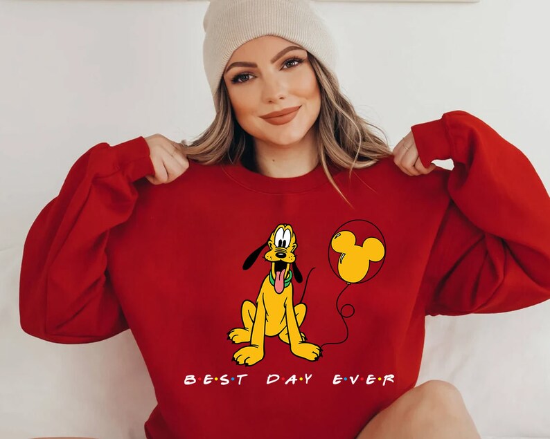 K&ouml;nnte beinhalten: Roter Pullover mit einem Cartoon-Bild von Pluto, der einen Mickey-Maus-Ballon h&auml;lt. Der Text "BEST DAY EVER" ist unter dem Bild gedruckt.